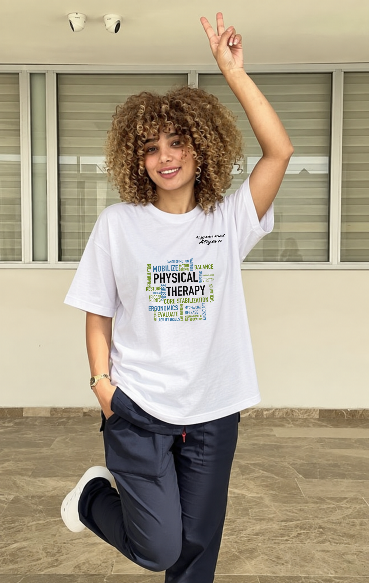 Fzt. Aliyeva beyaz PHYSICAL THERAPY ve siyah imza baskılı oversize tişörtle klinik ortamında, ön görünüm. PT-DSG-3820.