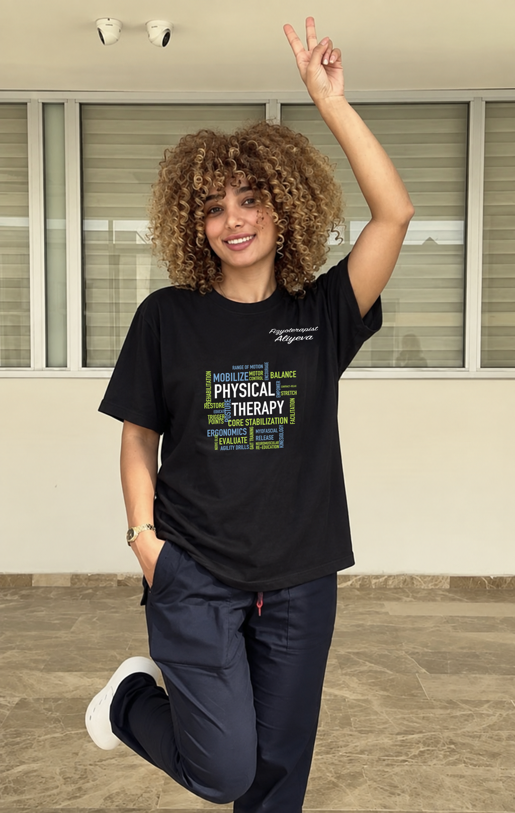 Fzt. Aliyeva siyah PHYSICAL THERAPY ve beyaz imza baskılı oversize tişörtle klinik ortamında, ön görünüm. PT-DSG-3821.