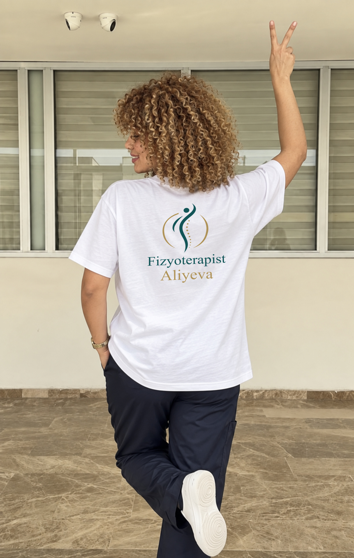 Fzt. Aliyeva büyük petrol yeşili ve altın logo baskılı beyaz oversize tişörtle klinik ortamında, sırtı dönük arka görünüm. PT-DSG-3823.